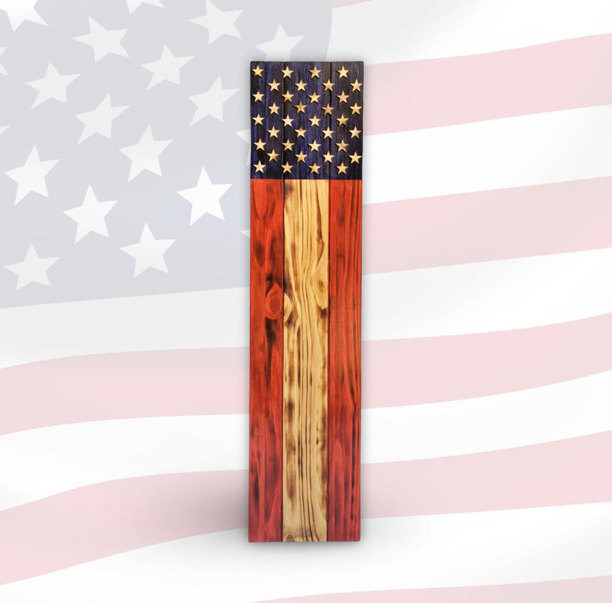 A handcrafted vertical wooden porch sign styled like the American flag, featuring multiple carved stars on a navy blue field with rustic red and natural woodgrain stripes, displayed against a faded flag backdrop.